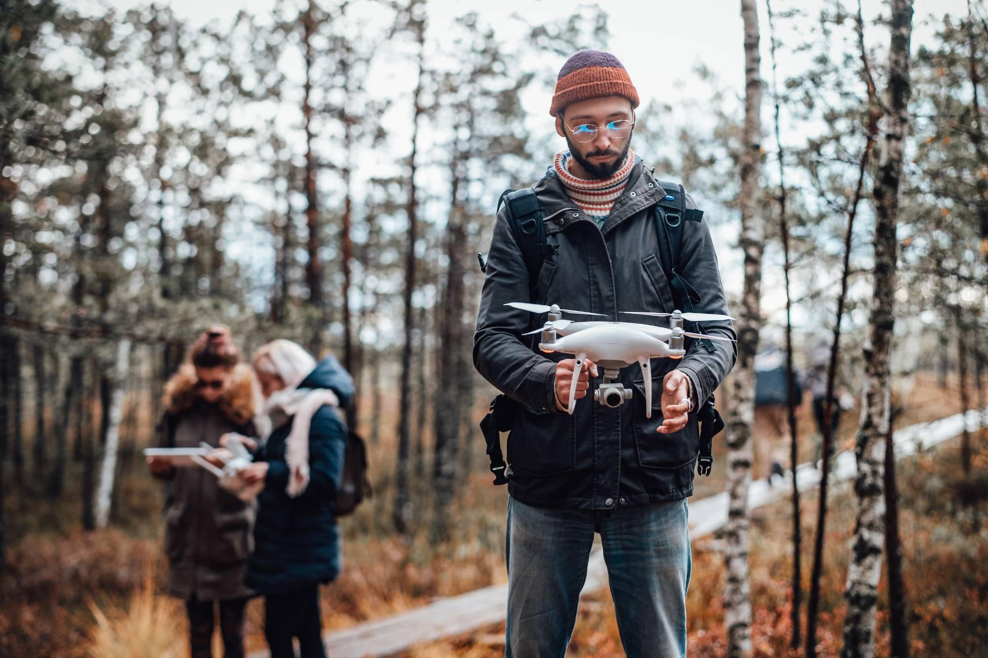 Bearded student with quadcopter in a wooded area with two others looking at terrain maps